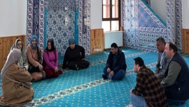 Bir caminin iç mekânında, turkuaz desenli halı üzerinde kadın ve erkeklerden oluşan bir grup daire şeklinde oturmuş. Duvarlar mavi-beyaz çinilerle süslü, sağda minber ve pencere görünüyor. Ortam sakin, sohbet ya da toplantı havası var.