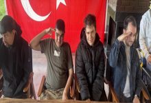 Türk bayrağı önünde yan yana oturan dört gençten ikisi asker selamı verirken gülümsüyor. Kapalı bir mekânda çekilen karede gençlerin önünde ahşap masa ve sandalyeler yer alıyor.