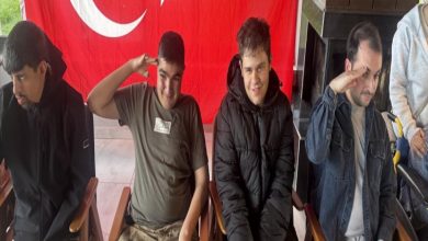 Türk bayrağı önünde yan yana oturan dört gençten ikisi asker selamı verirken gülümsüyor. Kapalı bir mekânda çekilen karede gençlerin önünde ahşap masa ve sandalyeler yer alıyor.