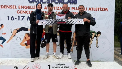 Bir spor müsabakası ödül töreni görülüyor. Üç basamaklı bir kürsü üzerinde dört kişi ayakta duruyor. Ortadaki sporcu birincilik basamağında ve boynunda madalya taşıyor. Grup, “ÖZALP GENÇLİK SK ŞAHİNLER” yazılı bir pankart tutuyor. Arkadaki afişte “Okul Sporları”, “Bedensel Engelliler Atletizm ve Puanlı Atletizm Gençlik Spor Şampiyonası” ifadeleri ve “16–17 Mayıs” tarihi yer alıyor. Arka planda sporcu illüstrasyonları bulunuyor. Atmosfer başarı ve gurur duygusu taşıyor.