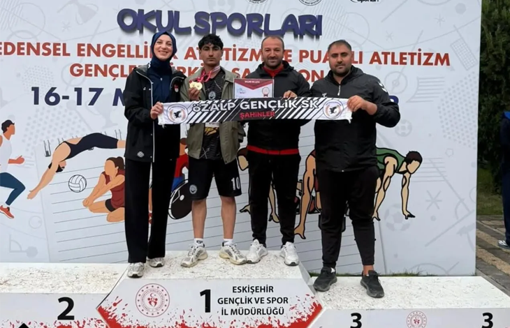 Bir spor müsabakası ödül töreni görülüyor. Üç basamaklı bir kürsü üzerinde dört kişi ayakta duruyor. Ortadaki sporcu birincilik basamağında ve boynunda madalya taşıyor. Grup, “ÖZALP GENÇLİK SK ŞAHİNLER” yazılı bir pankart tutuyor. Arkadaki afişte “Okul Sporları”, “Bedensel Engelliler Atletizm ve Puanlı Atletizm Gençlik Spor Şampiyonası” ifadeleri ve “16–17 Mayıs” tarihi yer alıyor. Arka planda sporcu illüstrasyonları bulunuyor. Atmosfer başarı ve gurur duygusu taşıyor.