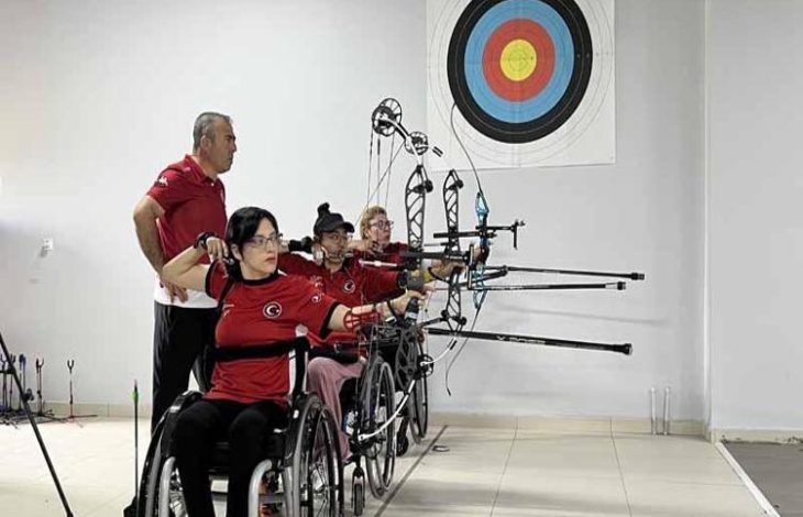 Gazi Oğuzhan Polat, kulüp antrenmanında ayakta durarak önünde bulunan tekerlekli sandalye kullanan okçuların atışını izliyor.