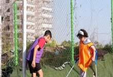 Fotoğrafta, iki çocuk açık havada bir futbol sahasında birlikte top oynarken görülüyor. Çocuklardan biri ampute ve koltuk değnekleriyle hareket ediyor; üzerinde turuncu bir yelek var. Diğer çocuk ise mor yelek giyiyor ve topa doğru yönelmiş durumda. Arka planda tel örgüler ve yüksek bir bina dikkat çekiyor.
