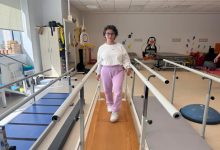 Fizik tedavi ve rehabilitasyon merkezinde genç bir kadın yürüme barları arasında yürüyüş egzersizi yapıyor. Spor kıyafetleriyle kameraya doğru ilerleyen kadın, dengeli şekilde adım atarken görülüyor. Ortamda çeşitli rehabilitasyon ekipmanları ve terapi alanları dikkat çekiyor.