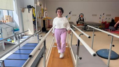 Fizik tedavi ve rehabilitasyon merkezinde genç bir kadın yürüme barları arasında yürüyüş egzersizi yapıyor. Spor kıyafetleriyle kameraya doğru ilerleyen kadın, dengeli şekilde adım atarken görülüyor. Ortamda çeşitli rehabilitasyon ekipmanları ve terapi alanları dikkat çekiyor.
