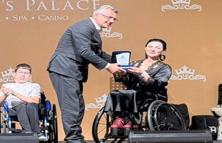 Sahnedeki törende, takım elbiseli bir erkek tekerlekli sandalyedeki kadına plaket takdim ediyor. Yan tarafta yine tekerlekli sandalyede oturan genç bir katılımcı görülürken, arka planda etkinliğin düzenlendiği salonun logosu yer alıyor. Fotoğraf, engelli bireylere yönelik farkındalık ve dayanışma temalı özel bir ödül gecesi atmosferini yansıtıyor.