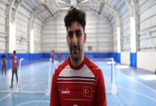 Kapalı bir spor salonunda, fileli bir sahada kırmızı-beyaz forma giymiş bir erkek sporcu kameraya dönük duruyor. Formasında Türk bayrağı bulunuyor. Arka planda aynı sahada antrenman yapan başka sporcular seçiliyor. Atmosfer antrenman ya da röportaj havasında.