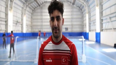 Kapalı bir spor salonunda, fileli bir sahada kırmızı-beyaz forma giymiş bir erkek sporcu kameraya dönük duruyor. Formasında Türk bayrağı bulunuyor. Arka planda aynı sahada antrenman yapan başka sporcular seçiliyor. Atmosfer antrenman ya da röportaj havasında.