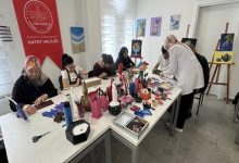 Bir atölye ortamında, kadınların bir masa etrafında oturup el işi ve sanat çalışmaları yaptığı, arka planda Hatay Valiliği yazılı kırmızı bir afişin bulunduğu bir sahne görülüyor.