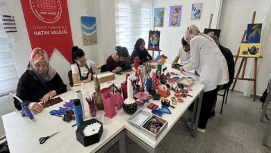 Bir atölye ortamında, kadınların bir masa etrafında oturup el işi ve sanat çalışmaları yaptığı, arka planda Hatay Valiliği yazılı kırmızı bir afişin bulunduğu bir sahne görülüyor.