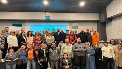 Bir konferans salonunda, farklı yaş gruplarından kalabalık bir grup sahne önünde toplu fotoğraf veriyor. Arka planda sunum ekranı yer alırken, katılımcılar yan yana dizilmiş. Aralarında tekerlekli sandalye kullanan bireyler de bulunuyor. Ortam, bir zirve ya da eğitim etkinliği sonrası çekilen hatıra fotoğrafı havası taşıyor.
