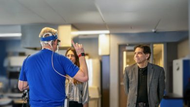 Kapalı bir laboratuvar ortamında başına elektronik cihaz takılı bir kişi el hareketiyle bir şey anlatırken, karşısında duran iki kişi dikkatle onu izliyor. Teknolojik ekipmanların bulunduğu sahne, bilimsel bir test ya da araştırma çalışmasını yansıtıyor.