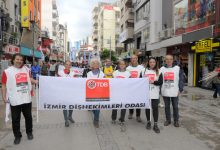 Bir grup insan, şehir merkezinde yürüyüş yaparken ön safta büyük bir pankart taşıyor. Pankartta “İzmir Dişhekimleri Odası” ve TDB logosu yer alıyor. Katılımcılar beyaz yelekler giymiş, arkada ise kalabalık bir kortej ilerliyor. Görüntü, örgütlü bir şekilde yapılan bir yürüyüş ve dayanışma etkinliğini yansıtıyor.