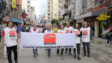 Bir grup insan, şehir merkezinde yürüyüş yaparken ön safta büyük bir pankart taşıyor. Pankartta “İzmir Dişhekimleri Odası” ve TDB logosu yer alıyor. Katılımcılar beyaz yelekler giymiş, arkada ise kalabalık bir kortej ilerliyor. Görüntü, örgütlü bir şekilde yapılan bir yürüyüş ve dayanışma etkinliğini yansıtıyor.