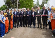 Açık alanda düzenlenen törende protokol üyeleri kurdele kesimi gerçekleştiriyor. Tören alanının iki yanında özel gereksinimli bireyler ve görevli personel yer alırken, arka planda sera çadırları ve ağaçlar görülüyor. Etkinlik resmi ve toplu katılımla gerçekleşiyor.
