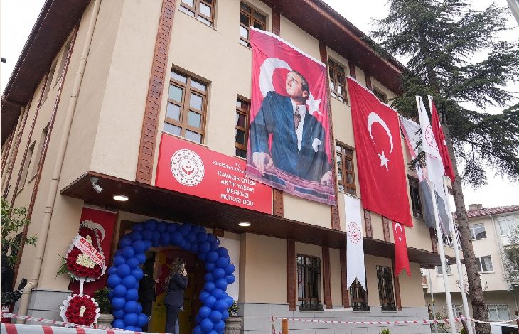 Bir kamu binasının dış cephesinde Türk bayrakları ve Mustafa Kemal Atatürk posterleri asılı görülüyor. Giriş kısmı mavi balonlarla süslenirken, binanın üzerinde “Kavacık Engelli Aktif Yaşam Merkezi Müdürlüğü” tabelası yer alıyor. Görsel, resmi açılış ya da etkinlik atmosferini yansıtıyor.