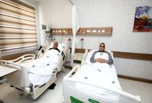 Bir hastane odasında, yan yana iki hasta yatağında yatan iki kişi görülüyor. Üzerlerinde hastane örtüleri var; biri dinlenir şekilde uzanırken diğeri ellerini göğsünde birleştirmiş. Oda temiz ve düzenli; başucunda tıbbi cihazlar ve duvarda ekipmanlar yer alıyor. Atmosfer sakin ve tedavi sürecini yansıtıyor.