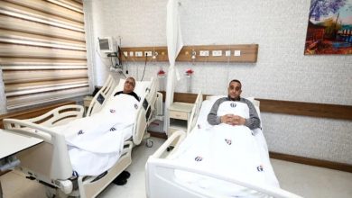 Bir hastane odasında, yan yana iki hasta yatağında yatan iki kişi görülüyor. Üzerlerinde hastane örtüleri var; biri dinlenir şekilde uzanırken diğeri ellerini göğsünde birleştirmiş. Oda temiz ve düzenli; başucunda tıbbi cihazlar ve duvarda ekipmanlar yer alıyor. Atmosfer sakin ve tedavi sürecini yansıtıyor.