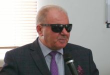 Yakın plan çekilen fotoğrafta, takım elbise ve mor kravat takan yaşlı bir erkek elinde mikrofonla konuşuyor. Siyah güneş gözlüğü kullanan kişinin arka planında pencere ve iç mekân detayları görülüyor. Görsel, görme engelli farkındalığı üzerine yapılan bir röportaj ya da konuşma anını yansıtıyor.