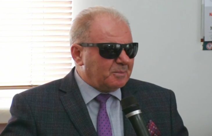 Yakın plan çekilen fotoğrafta, takım elbise ve mor kravat takan yaşlı bir erkek elinde mikrofonla konuşuyor. Siyah güneş gözlüğü kullanan kişinin arka planında pencere ve iç mekân detayları görülüyor. Görsel, görme engelli farkındalığı üzerine yapılan bir röportaj ya da konuşma anını yansıtıyor.