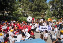 Kalabalık bir yürüyüşte, renkli şapkalar takan çocuklar ve yetişkinler pankartlar taşıyarak büyük bir panda maskotunun etrafında toplanmış şekilde görülüyor.