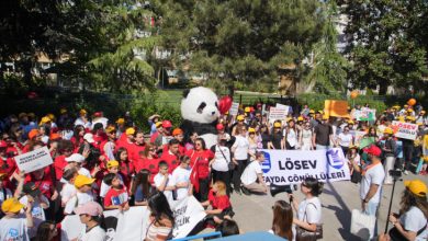 Kalabalık bir yürüyüşte, renkli şapkalar takan çocuklar ve yetişkinler pankartlar taşıyarak büyük bir panda maskotunun etrafında toplanmış şekilde görülüyor.