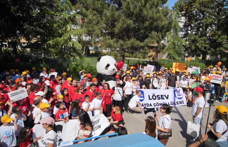 Kalabalık bir yürüyüşte, renkli şapkalar takan çocuklar ve yetişkinler pankartlar taşıyarak büyük bir panda maskotunun etrafında toplanmış şekilde görülüyor.