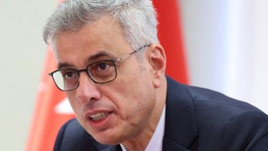 Bakan Memişoğlu konuşma yaparken görülüyor. Arka planda kırmızı‑beyaz bir bayrak bulunuyor; sahne resmi ve ciddi bir ortam hissi veriyor.