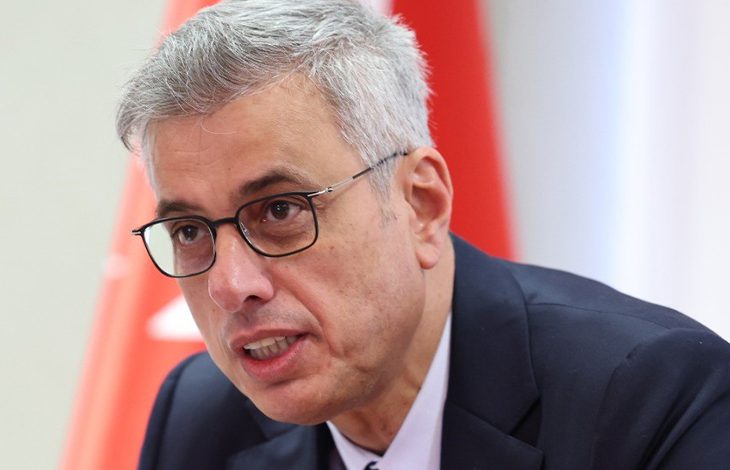 Bakan Memişoğlu konuşma yaparken görülüyor. Arka planda kırmızı‑beyaz bir bayrak bulunuyor; sahne resmi ve ciddi bir ortam hissi veriyor.