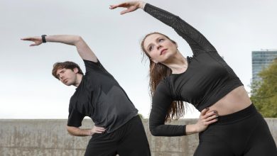 Açık havada, spor kıyafetli bir kadın ve bir erkek yanlara doğru esneme hareketi yapıyor. Kolları yukarı uzanmış, gövdeleri yana eğilmiş. Arka planda sade bir gökyüzü ve uzakta bir bina görünüyor. Atmosfer egzersiz ve sağlıklı yaşam odaklı.