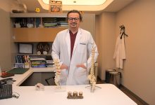 Beyaz önlük giymiş Op. Dr. Bülent Bozyiğit, bir muayenehane ya da klinik ofisinde ayakta duruyor. Önündeki masada omurga ve omur modelleri bulunuyor. Arka planda kitaplar, klasörler ve sertifikalarla dolu düzenli bir raf sistemi yer alıyor. Ortam profesyonel, temiz ve tıbbi bir çalışma alanını yansıtıyor.