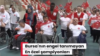 Kapalı spor salonunda düzenlenen etkinlikte tekerlekli sandalye kullanan bireyler ve gönüllüler Türk bayraklarıyla yürüyüş yapıyor. Katılımcıların büyük bölümü beyaz tişört ve kırmızı yelekler giyerken, etkinlik coşkulu ve dayanışma dolu bir atmosfer yansıtıyor.