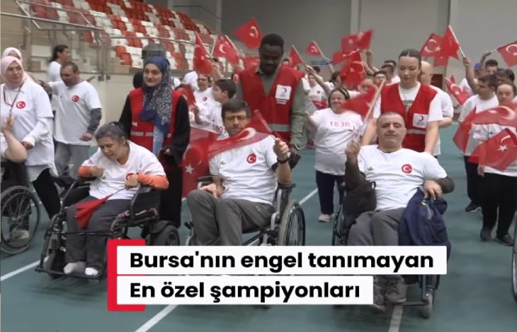 Kapalı spor salonunda düzenlenen etkinlikte tekerlekli sandalye kullanan bireyler ve gönüllüler Türk bayraklarıyla yürüyüş yapıyor. Katılımcıların büyük bölümü beyaz tişört ve kırmızı yelekler giyerken, etkinlik coşkulu ve dayanışma dolu bir atmosfer yansıtıyor.