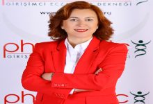 Görselde, kırmızı bir takım elbise ve beyaz gömlek giyen bir kadın kamera karşısında poz veriyor. Kollarını çapraz şekilde bağlamış, gülümseyen bir ifadeye sahip. Arka planda bir derneğe ait olduğu anlaşılan logo ve yazılar bulunuyor.