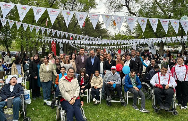 Ağaçlık bir park alanında, üzerlerinde "Hayal Etmek Özgürlüktür" yazılı flamaların asılı olduğu bir meydanda; tekerlekli sandalyedeki bireyler, çocuklar ve yetişkinlerin bir arada olduğu kalabalık bir grup fotoğrafıdır. Sosyal dayanışma temalı bir açık hava etkinliğini yansıtmaktadır.