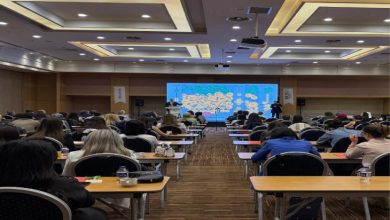 Geniş bir konferans salonunda düzenlenen seminerde katılımcılar sıralı masalarda oturarak sunumu dinliyor. Salonun ön kısmındaki büyük ekranda sunum yansıtılırken, konuşmacılar sahnede yer alıyor. Etkinlik yoğun katılımla gerçekleşiyor.