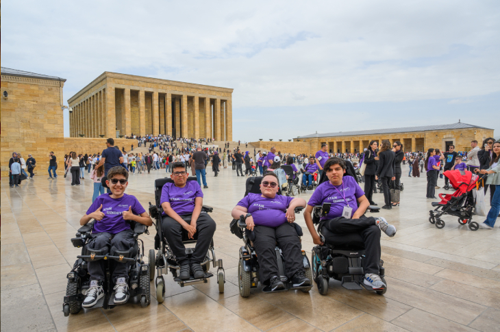 Anıtkabir avlusunda, akülü tekerlekli sandalyelerinde yan yana poz veren mor tişörtlü dört SMA'lı gencin yer aldığı bir karedir. Gençlerin tişörtlerinde "Atam İzindeyiz" ifadesi yer alırken, arka planda kalabalık bir ziyaretçi grubu ve Anıtkabir Mozolesi görülmektedir.