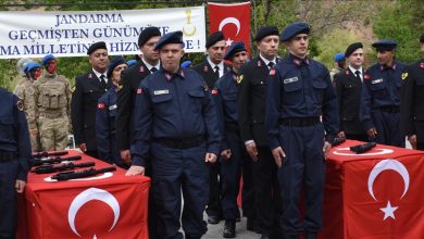 Bir grup jandarma personeli tören düzeninde duruyor; önlerindeki masalarda Türk bayrakları ve sergilenen tabancalar, arkada ise “Jandarma – Daima Milletin Hizmetinde” yazılı büyük bir afiş bulunuyor.