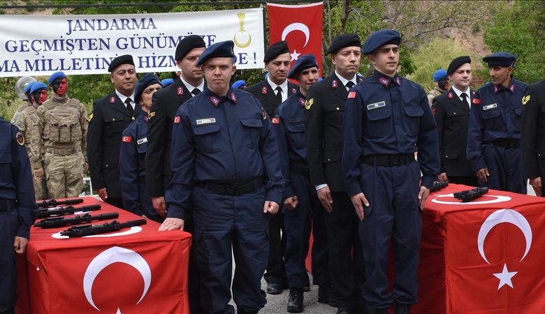 Bir grup jandarma personeli tören düzeninde duruyor; önlerindeki masalarda Türk bayrakları ve sergilenen tabancalar, arkada ise “Jandarma – Daima Milletin Hizmetinde” yazılı büyük bir afiş bulunuyor.
