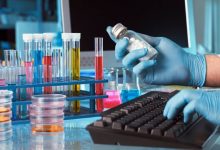 Laboratuvar ortamında mavi eldivenli bir kişinin, renkli kimyasal sıvılarla dolu deney tüplerinin yanında beyaz tabletler içeren bir şişeyi incelerken bilgisayar klavyesini kullandığı görülüyor.