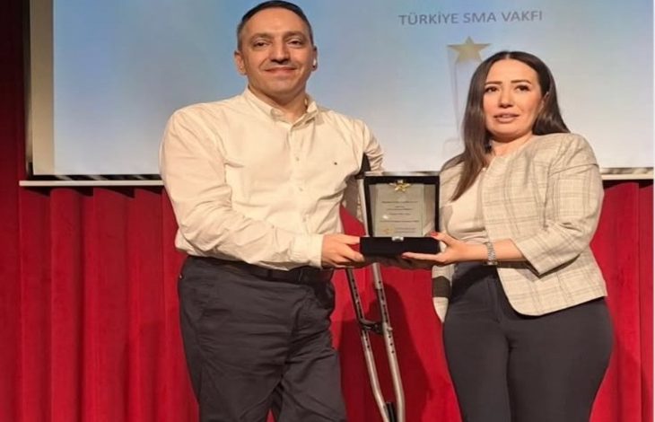 Görselde, Altın Dokunuş Ödülleri töreninde bir kadın ve bir erkek sahne önünde birlikte ödül plaketi tutarken görülmektedir. Arka planda kırmızı perdeler ve üzerinde "Türkiye SMA Vakfı" yazılı bir yansıtma ekranı bulunmaktadır. Ödülü verenin yanında bir koltuk değneği dikkat çekmektedir.