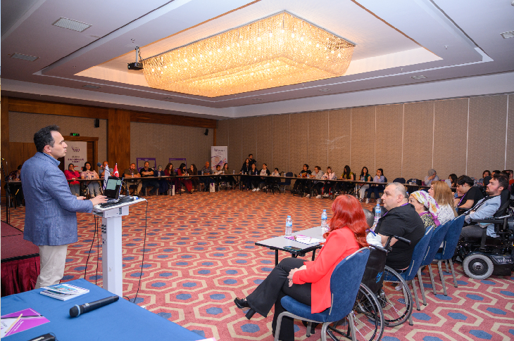 Bir konferans salonunda düzenlenen toplantıda konuşmacı kürsüden katılımcılara sunum yapıyor. Salonda farklı yaş gruplarından bireyler, aileler ve tekerlekli sandalye kullanan katılımcılar yer alırken, etkinlik dayanışma, gönüllülük ve toplumsal farkındalık atmosferi taşıyor.