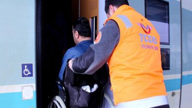 Turuncu yelekli bir görevli, tekerlekli sandalye kullanan bir yolcunun trene binişine yardımcı oluyor. Tren kapısının yanında engelli erişim işareti yer alırken, görüntü erişilebilir ulaşım hizmetlerini yansıtıyor.