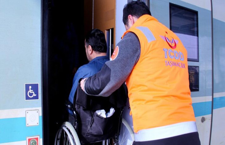 Turuncu yelekli bir görevli, tekerlekli sandalye kullanan bir yolcunun trene binişine yardımcı oluyor. Tren kapısının yanında engelli erişim işareti yer alırken, görüntü erişilebilir ulaşım hizmetlerini yansıtıyor.