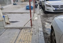 Dar bir şehir sokağında, kaldırım üzerindeki inşaat çalışmaları ve düzensiz park edilmiş araçlar nedeniyle yaya geçişi zorlaşırken, sarı hissedilebilir yüzeyin önünde metal direkler ve molozlar dikkat çekiyor.