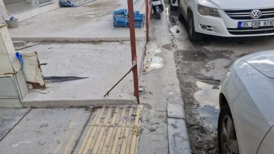 Dar bir şehir sokağında, kaldırım üzerindeki inşaat çalışmaları ve düzensiz park edilmiş araçlar nedeniyle yaya geçişi zorlaşırken, sarı hissedilebilir yüzeyin önünde metal direkler ve molozlar dikkat çekiyor.