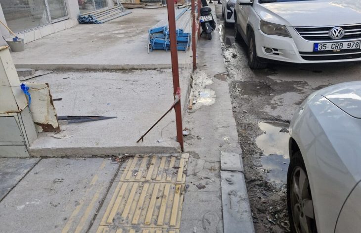 Dar bir şehir sokağında, kaldırım üzerindeki inşaat çalışmaları ve düzensiz park edilmiş araçlar nedeniyle yaya geçişi zorlaşırken, sarı hissedilebilir yüzeyin önünde metal direkler ve molozlar dikkat çekiyor.
