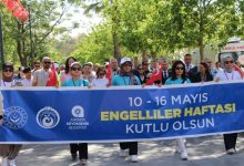 Kalabalık bir grup, ellerinde Türk bayraklarıyla “10–16 Mayıs Engelliler Haftası Kutlu Olsun” yazılı büyük bir pankart taşıyarak ağaçlarla çevrili bir alanda yürüyüş yapıyor.