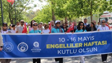 Kalabalık bir grup, ellerinde Türk bayraklarıyla “10–16 Mayıs Engelliler Haftası Kutlu Olsun” yazılı büyük bir pankart taşıyarak ağaçlarla çevrili bir alanda yürüyüş yapıyor.
