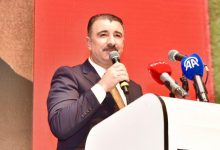 Bir kürsüde konuşma yapan takım elbiseli bir kişi elindeki mikrofonla kalabalığa hitap ediyor. Kürsü üzerinde çeşitli basın kuruluşlarına ait mikrofonlar bulunurken, arka plandaki kırmızı ekran etkinliğe resmi bir atmosfer katıyor. Görsel, bir basın açıklaması veya toplu konuşma anını yansıtıyor.